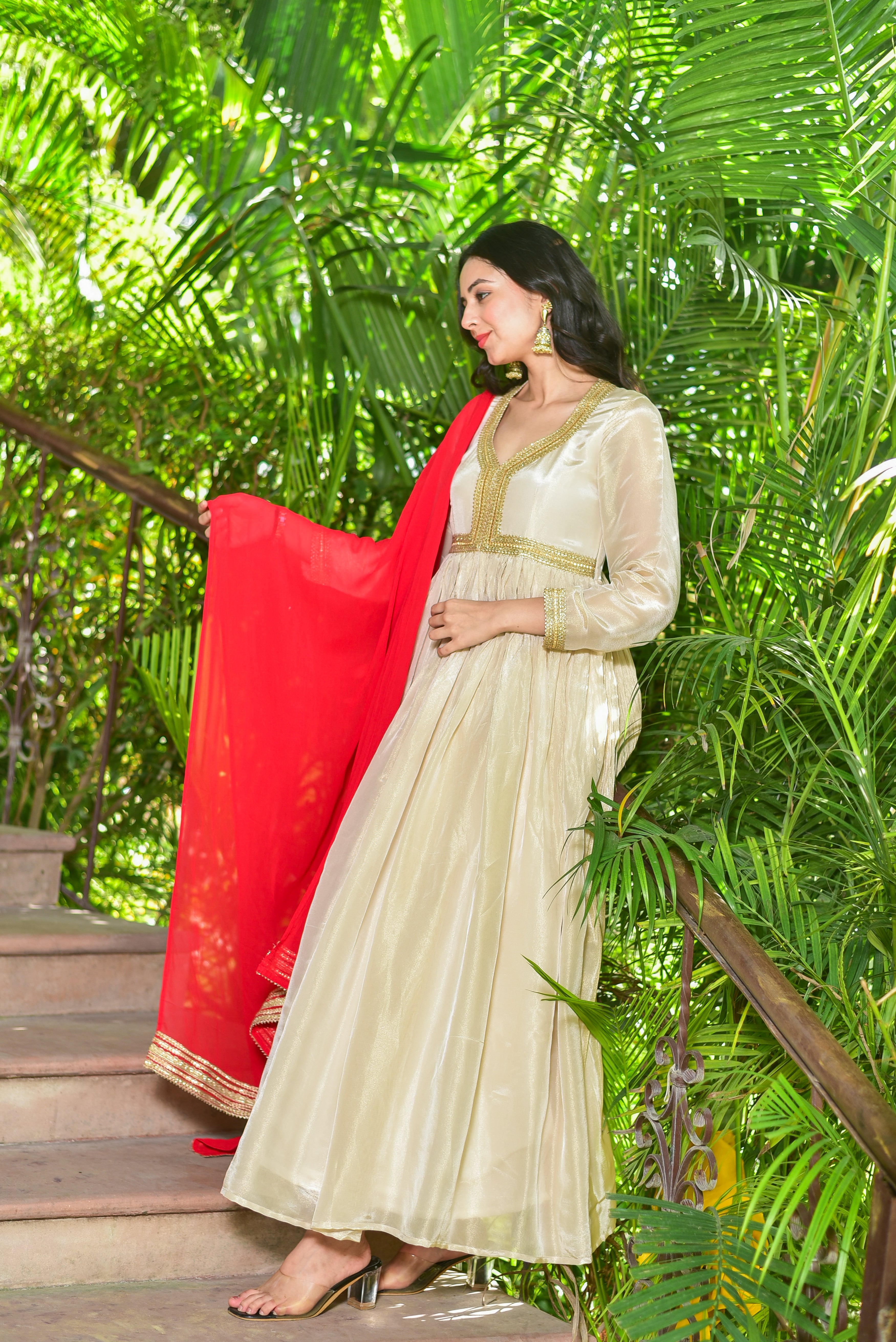 Woman in a cream-colored dress with a red dupatta standing on steps surrounded by green foliage.